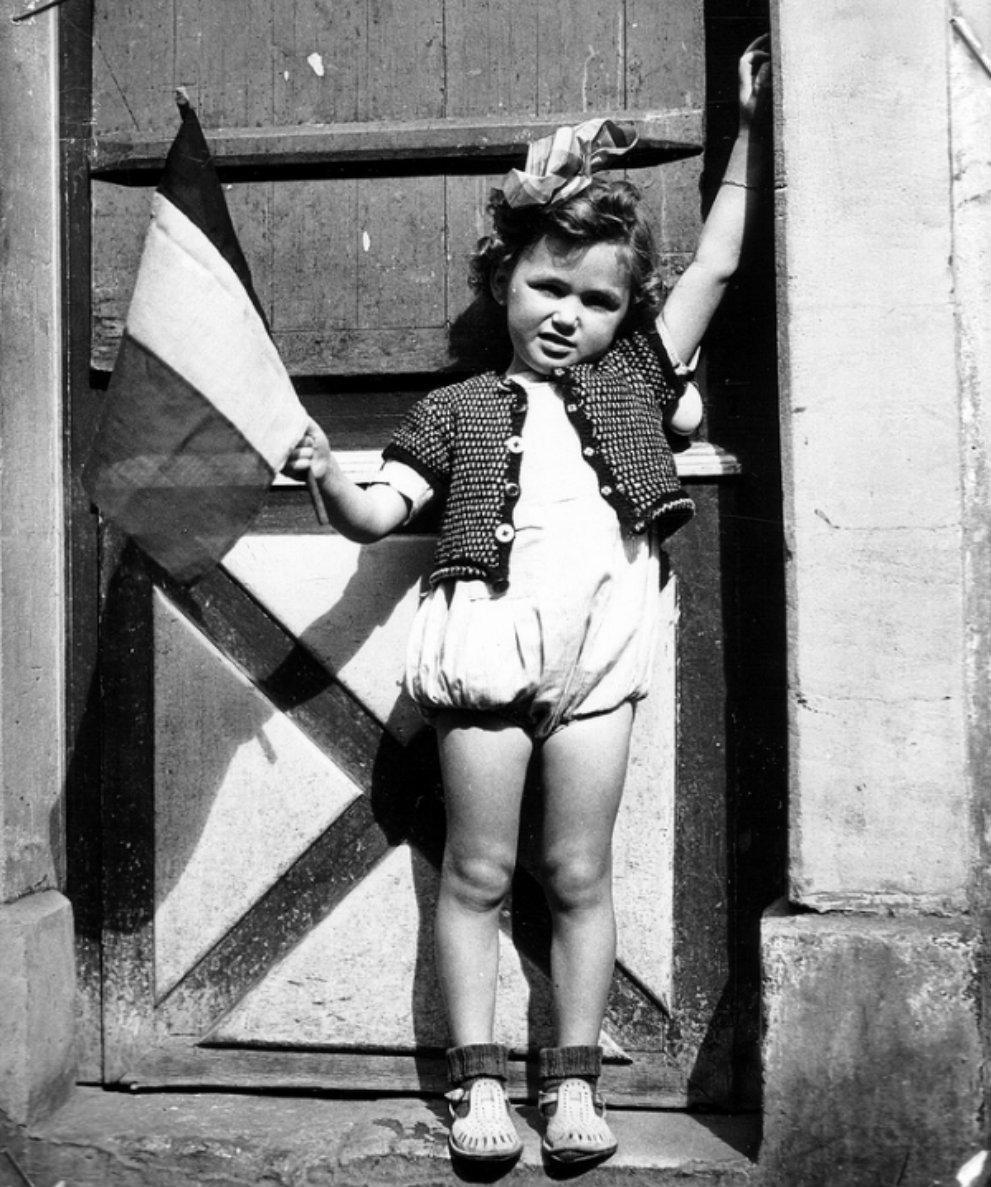 A little French girl stands in the doorway waves the French flag after liberation of her town by the British. Port-en-Bessin-Huppain, Calvados, Lower Normandy, France. July 19.jpg